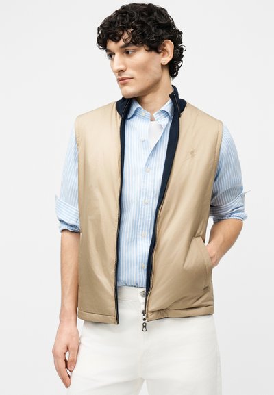 Νεαρός άνδρας που φοράει μπεζ γιλέκο με φερμουάρ πάνω από μπλε ριγέ πουκάμισο με μανίκια που έχουν γυριστεί και άσπρο παντελόνι, κοιτάζει προς τα αριστερά του.