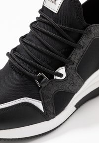 Černá sportovní bota s texturovanými šedými akcenty, kulatými tkaničkami a bílou podrážkou. Materiály zahrnují syntetickou tkaninu a kůži.