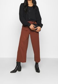 Černá halenka s dlouhým rukávem, knoflíky a balónovými rukávy v kombinaci se středně vysokými hnědými širokými kalhotami. Obuv tvoří černé kotníkové boty.