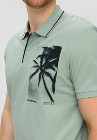 Jasnozielona koszulka polo z czarnymi akcentami, z graficznym motywem palmy na klatce piersiowej. Wykonana z bawełny, z krótkim rękawem i klasycznym kołnierzykiem.