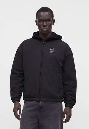 Homme portant une veste noire à capuche zippée avec le logo EA7 sur le côté gauche de la poitrine et un jean noir délavé, debout sur un fond blanc uni.