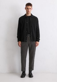 Black ribbed cardigan with button closure, worn over a black shirt, paired with dark gray corduroy pants and black lace-up shoes.