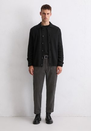 Zwarte geribbelde cardigan met knoopsluiting, gedragen over een zwart shirt, gecombineerd met donkergrijze corduroy broek en zwarte veterschoenen.