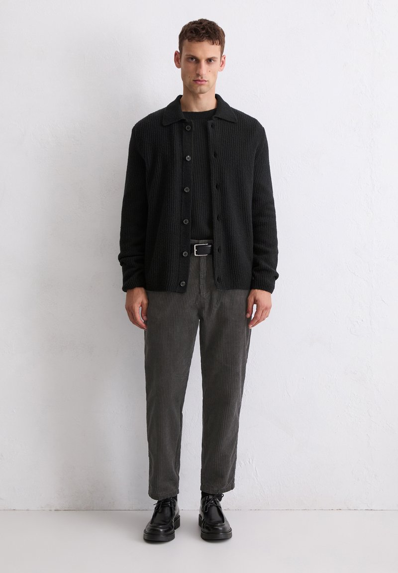 Black ribbed cardigan with button closure, worn over a black shirt, paired with dark gray corduroy pants and black lace-up shoes.