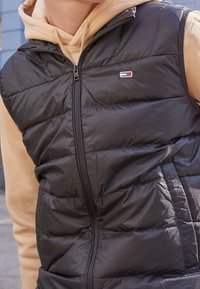 Czarna kamizelka puchowa z zapięciem na zamek, z poziomymi przeszyciami i małą naszywką z logo. Noszona na beżowym hoodie.