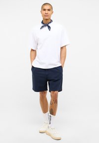 Camiseta blanca de algodón, pantalones cortos azul marino, bandana azul con estampado y zapatillas blancas con suelas beige. Presenta tatuajes visibles en la pierna izquierda.