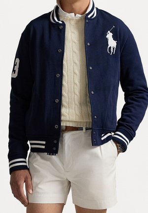 Homme portant une veste universitaire bleu marine avec des rayures blanches, un pull en maille torsadée crème, un short blanc et une montre au poignet gauche.