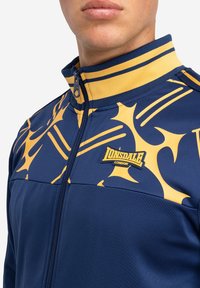 Veste marine avec des motifs abstraits jaunes, col haut et fermeture éclair. Présente le logo Lonsdale à l'avant. Texture de tissu lisse.