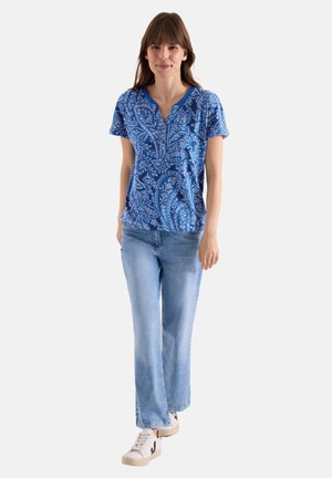 Femme marchant vers l’avant portant un haut bleu à manches courtes imprimé paisley, jean bleu clair et baskets blanches sur fond uni.