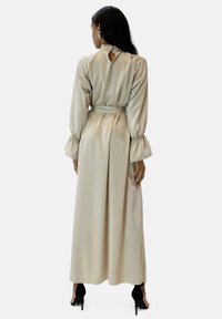 Lang beige satin kjole med høj hals, puffede ærmer og åben ryg med nøglehul. Har bundet talje og flydende silhuet. Sorte hæle båret.