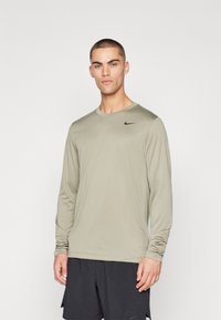 Langärmliges, olivgrünes Sportshirt aus glattem Material, mit einem schwarzen Nike-Logo auf der Brust, kombiniert mit schwarzen Shorts.