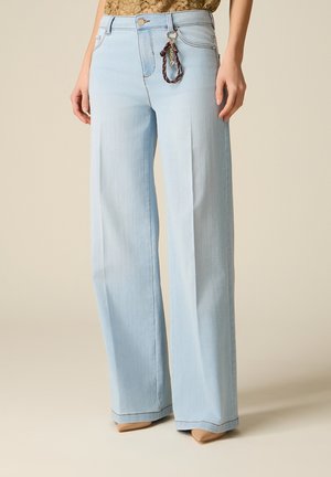 Femme portant un jean large bleu clair avec un porte-clés tressé marron attaché à la boucle de ceinture avant et des chaussures beiges à bout pointu.