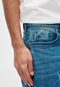 Μπλε τζην παντελόνι με ανοιχτό πλύσιμο, με καμπυλωτή μπροστινή τσέπη και ένα μικρό, μπλε κεντημένο λογότυπο στην τσέπη. Χέρι resting πάνω στη μέση.