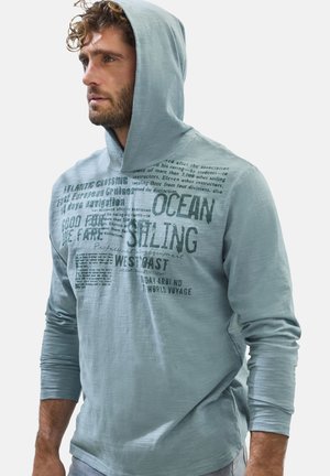 Mann mit lockigem Haar trägt einen hellblauen Kapuzenpullover mit schwarzem Seglertext auf der Vorderseite und schaut nach links.