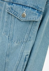 Světle modrá džínová bunda s špicatým límcem, knoflíkovým zapínáním a náprsní kapsou s klopou. Viditelné výrazné prošívání.