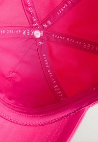 Intérieur d'une casquette en tissu rose vif avec des bandes de soutien croisées portant l'inscription "BAKER BY TED BAKER" et un bouton central.