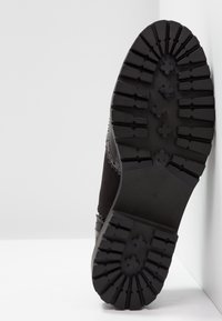 Semelle de chaussure noire avec des crampons profonds et dessinés, appuyée verticalement contre un mur d'angle blanc.