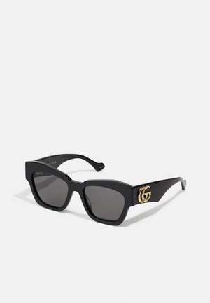 Gafas de sol rectangulares negras con lentes oscuros, que presentan el logo GG en oro en el costado. Fabricadas de plástico brillante con patillas rectas.
