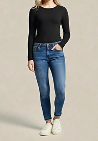 Femme debout avec une main dans la poche, portant un haut noir à manches longues, un jean bleu et des baskets blanches sur un fond beige.