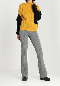 Žlutý texturovaný pletený svetr s černými dlouhými rukávy, doplněný o vzorované rozšířené kalhoty. Černé kotníkové boty dokončují outfit.