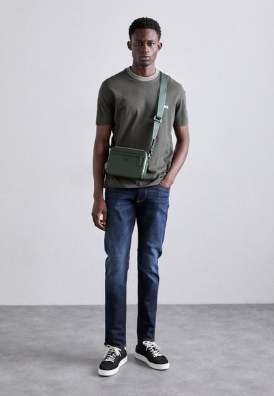 Grønn crossover-veske med justerbar stropp, brukt over en mørk grønn t-skjorte og mørk denim jeans, med svarte sneakers med hvite såler.