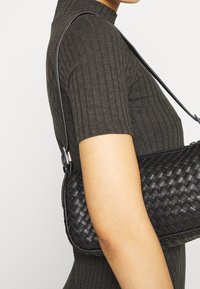 Svart vävd läderhandväska med slät finish och justerbar rem, buren under armen. Har en elegant, kompakt rektangulär form.