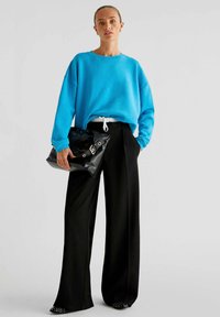 Blauwe oversized sweatshirt, zwarte wijde broek en een glanzende zwarte handtas met zilveren accenten. De outfit heeft een trekkoord in de taille.