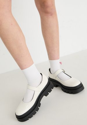 Scarpe Mary Jane in vernice bianca con una suola nera spessa, dotate di una cinghia sopra il collo del piede e indossate con calze bianche.