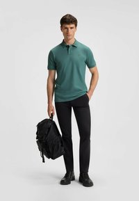 Polo di colore verde acqua con maniche corte e un piccolo logo sul petto, abbinato a pantaloni neri e uno zaino nero. Il modello è in piedi con un'espressione neutra.