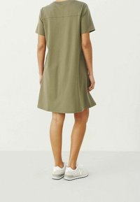 Vestido de algodón verde oliva con mangas cortas, corte relajado y forma en línea A, combinado con zapatillas gris claro. Textura suave y diseño minimalista.