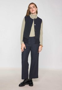 Gilet a maglia navy con frange, chiusura a bottoni; abbinato a un dolcevita a coste verde chiaro e pantaloni a gamba larga scuri. Scarpe nere stringate.