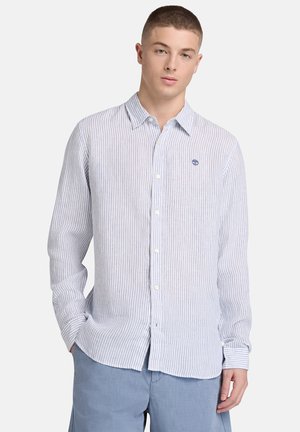 Giovane uomo con camicia a righe bianche e azzurre e pantaloni blu, in piedi con una mano in tasca contro uno sfondo semplice.