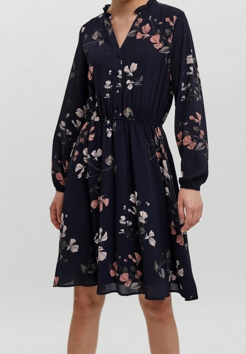 Femme portant une robe bleu marine mi-longue avec manches longues et motifs floraux roses et blancs, dotée d'une taille cintrée et d'un col boutonné.