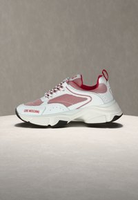 Λευκό και κόκκινο sneaker Love Moschino με πάνελ από δίχτυ, παχύ πάτο και κορδόνια με σχέδιο, σε ουδέτερη επιφάνεια και φόντο.