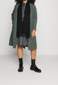 Grön ullkappa över en svart klänning, i kombination med en svart fransad scarf och blanka svarta ankelskor. Släta texturer, oversized passform, lager på lager-look.