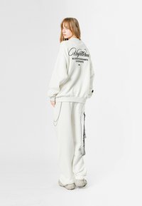 Sudadera blanca de gran tamaño con texto negro en la parte posterior, combinada con pantalones de chándal a juego. Presenta un detalle de cadena plateada en los pantalones.