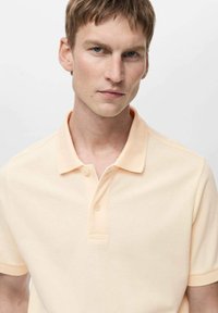 Světle broskvová polo košile vyrobená z měkké, texturované látky. Obsahuje klasický límeček, dva knoflíky a krátké rukávy.