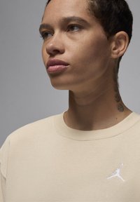 Felpa beige con scollo tondo e dettagli a costine. Presenta un logo Jumpman bianco ricamato sul petto sinistro. Persona con capelli corti.