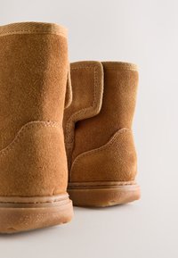 Cizme maro din piele intoarsa cu o suprafață texturată, formă rotunjită și talpă din cauciuc. Prezintă o margine din țesătură bej contrastantă de-a lungul marginii superioare.