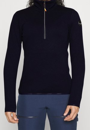 Kvinde iført en sort tekstureret langærmet pullover med halv lynlås og blå bukser med lynlåslommer.