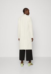 Palton lung, cu tăietură dreaptă, de culoare albă, realizat dintr-un material neted. Are mâneci lungi, collar cu vârf și un design minimalist. Pantaloni negri cu picioare largi și sandale verzi cu toc bloc completează ținuta.