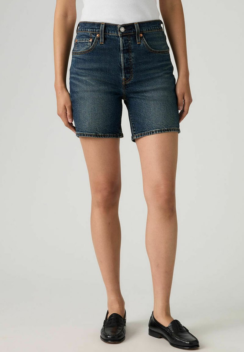 Shorts en denim bleu à coupe droite, dotés de poches avant, passants pour ceinture et d'un ourlet brut. Associés à des mocassins noirs.