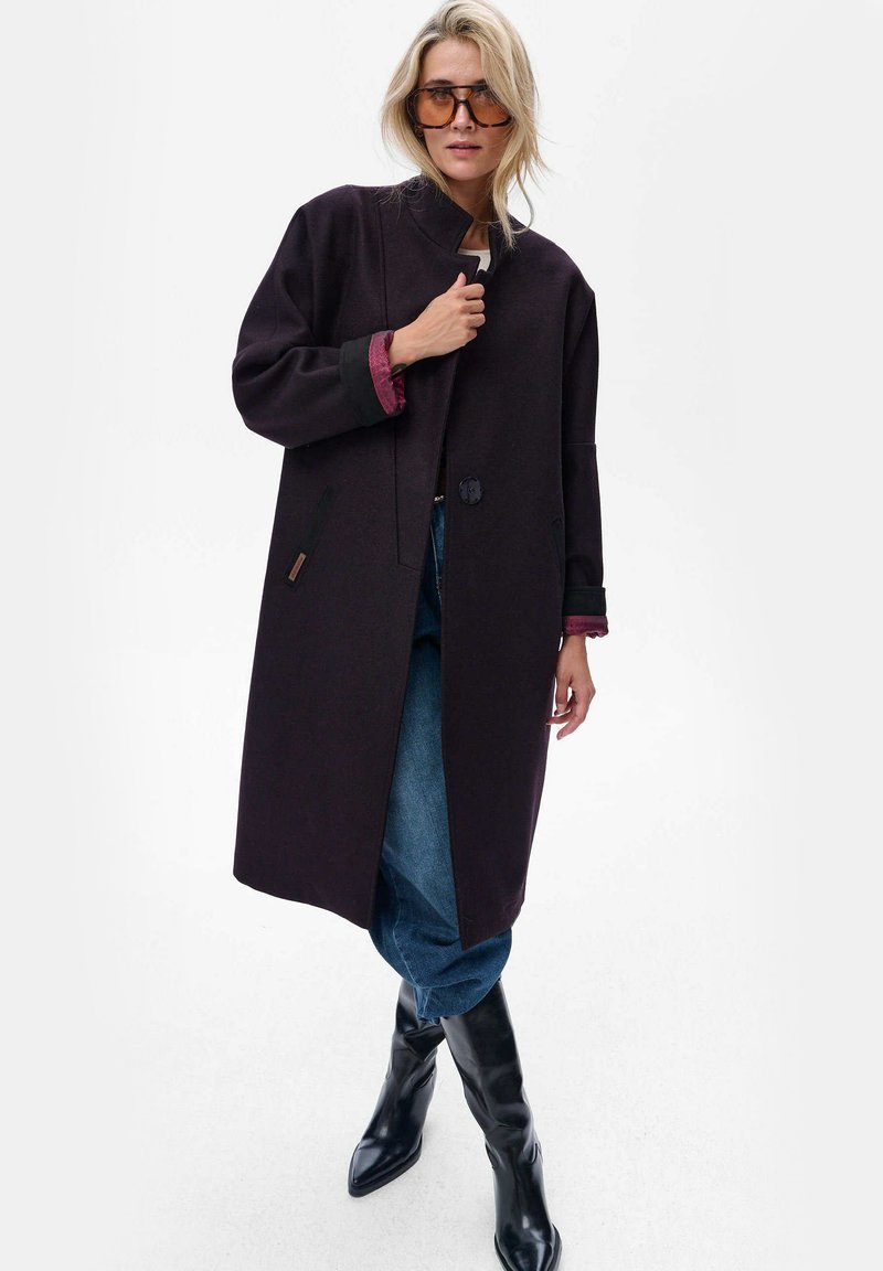 Lange, donkerviolet jas met een hoge kraag, knoopsluiting en contrasterende mouwen. Draagt over een blauwe jeans en zwarte laarzen tot de knie.