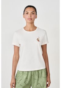 Camiseta de algodón crema con mangas cortas enrolladas, que presenta un colorido diseño bordado en el pecho. Combinada con pantalones cortos verdes con textura.