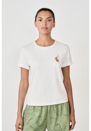 Camiseta de algodón crema con mangas cortas enrolladas, que presenta un colorido diseño bordado en el pecho. Combinada con pantalones cortos verdes con textura.