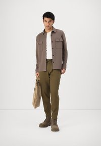Camisa castanha de botão com dois bolsos no peito, t-shirt branca por baixo, calças verde-azeitona, mala de mão bege e botas castanhas de atacadores.