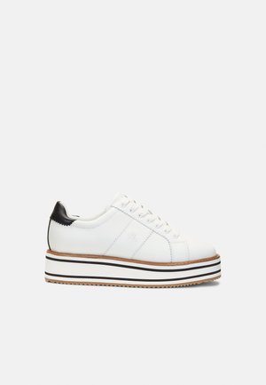 Hvid platform-sneaker med sort hælaccen, snørebånd foran og stribet sål i hvide, sorte og brune farver på en ensfarvet baggrund.