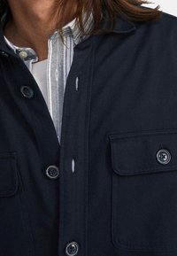 Närbild av en person som bär en marinblå knäppt jacka över en vit och grå randig skjorta med krage, där knappar och bröstficka syns.