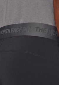 Juodi sportiniai šortai su lygiu paviršiumi ir elastinga kilpine juosta, ant kurios matomas pilkas „The North Face“ logotipas. Matoma sustiprinta siūlė.