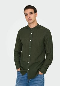 Chemise verte à manches longues avec un col mandarin, dotée d'une fermeture à boutons et de boutons blancs. Fabriquée en tissu texturé, elle est associée à un jean bleu.